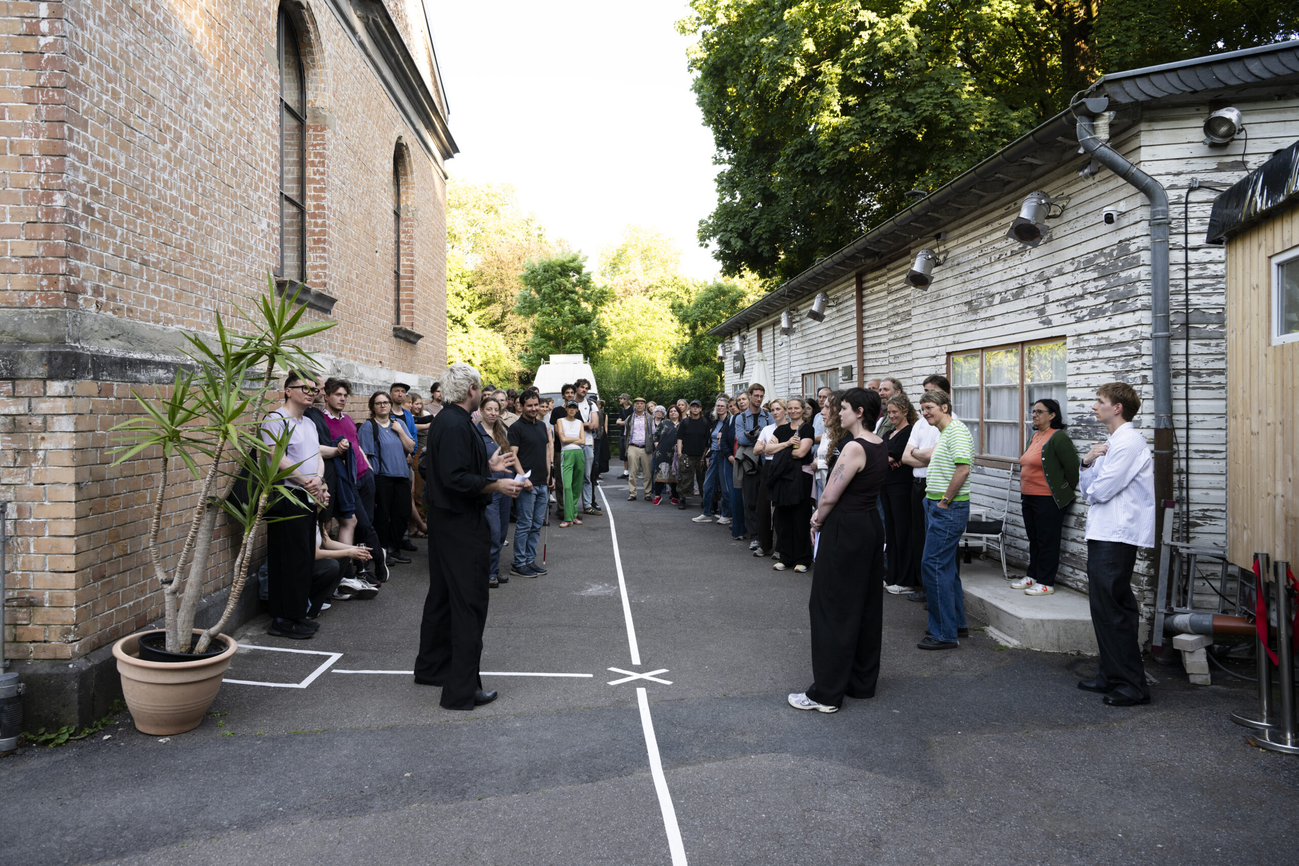 Eine Große Gruppe von Menschen steht draußen zwischen zwei Gebäuden (Backstein links, verwitterte Holzfassade rechts) in einem mit weißen Linien markierten Hof und hört einer männlich gelesenen Person zu, die in der Mitte des markierten Bereichs steht.