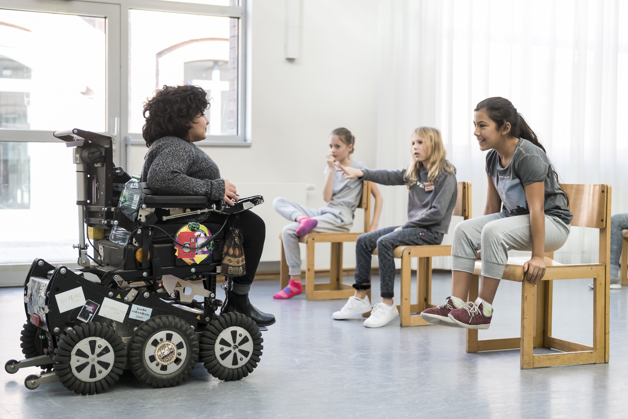 Eine weiblich gelesene Person in einem elektrischen Rollstuhl mit Geländefahrwerk unterhält sich mit drei Kindern, die auf Holzstühlen sitzen, in einem hellen Raum mit großen Fenstern.