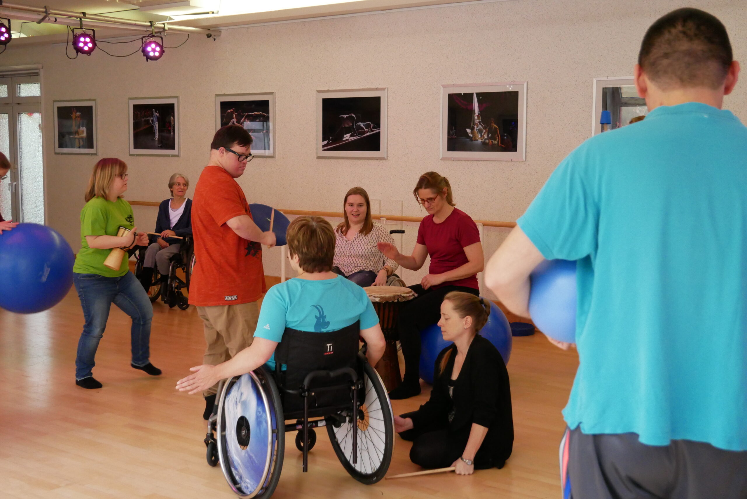 Gruppe von Personen musiziert gemeinsam mit Trommeln und blauen Bällen in einem Tanzsaal, darunter eine Person im Rollstuhl und eine Person, die kniend eine Trommel spielt. An der Wand hängen gerahmte Fotografien.