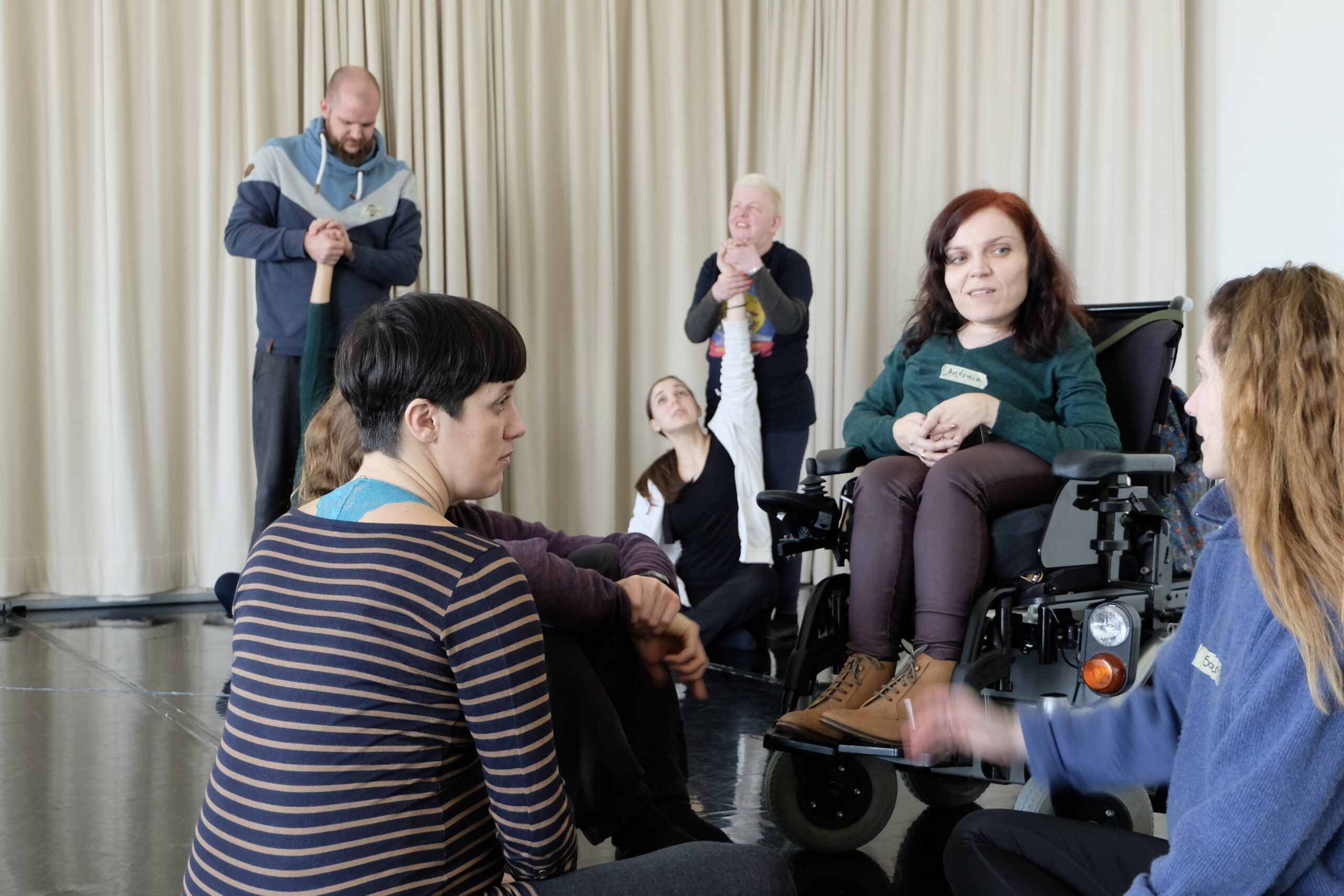 Gruppe von sieben Personen in einem Raum mit beigem Vorhang, teils sitzend, teils stehend mit verschränkten erhobenen Armen. Zwei Personen sitzen in Elektrorollstühlen, eine trägt ein Namensschild „Antonia".