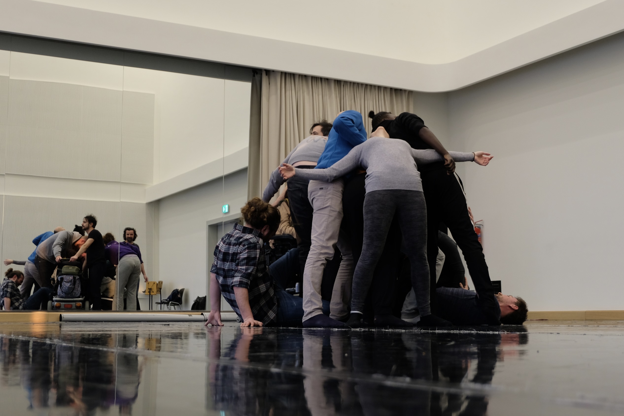 Gruppe von Tänzer:innen, die sich eng umschlungen zu einer Art menschlichem Knäuel formen, mit ausgestreckten Armen, in einem Spiegelsaal mit Vorhang. Eine Person liegt am Boden, eine weitere kriecht heran.