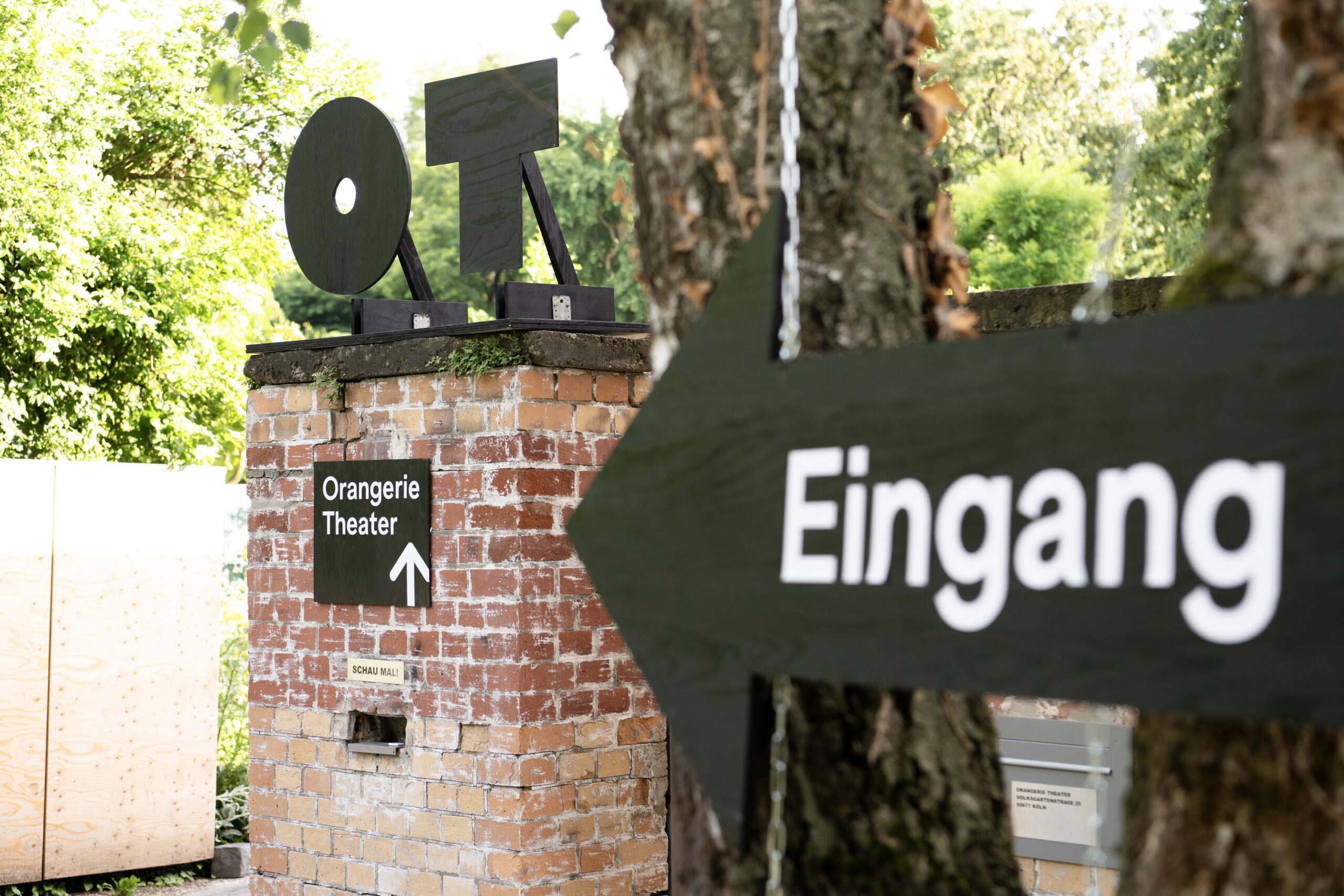 Nahaufnahme eines Wegweisers am Eingang eines Backsteingebäudes im Grünen. Auf der Mauer steht ein Holzlogo mit den Buchstaben „OT", darunter ein Schild „Orangerie Theater" mit Pfeil nach oben. Im Vordergrund unscharf ein Wegweiser mit der Aufschrift „Eingang".