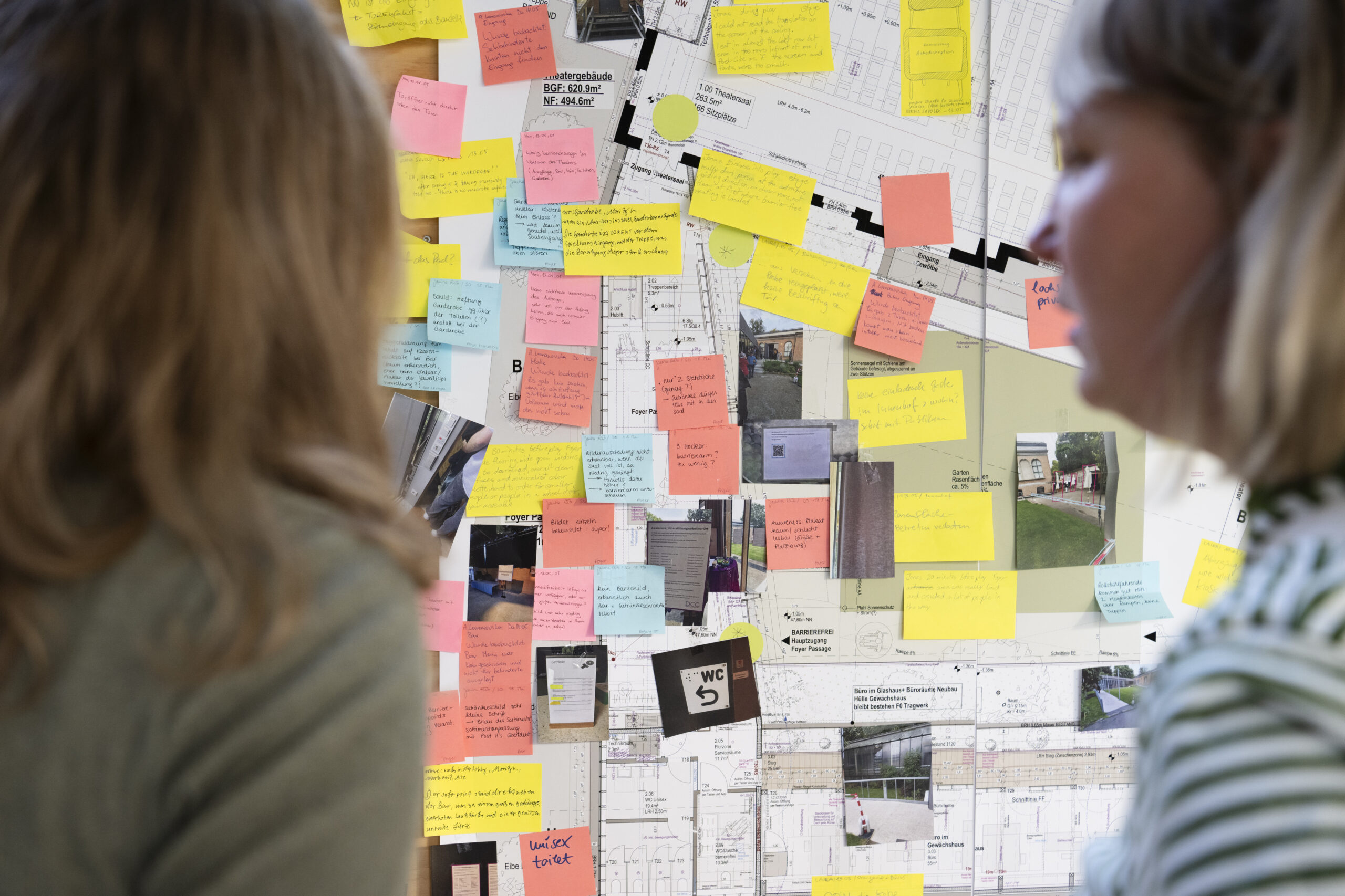 wei Personen betrachten von rechts und links eine große Pinnwand mit Grundrissplänen eines Theatergebäudes, die dicht mit bunten Klebezetteln (gelb, rosa, orange, blau) und Fotos beklebt ist – offenbar Feedback- und Planungsnotizen zu Barrierefreiheit.