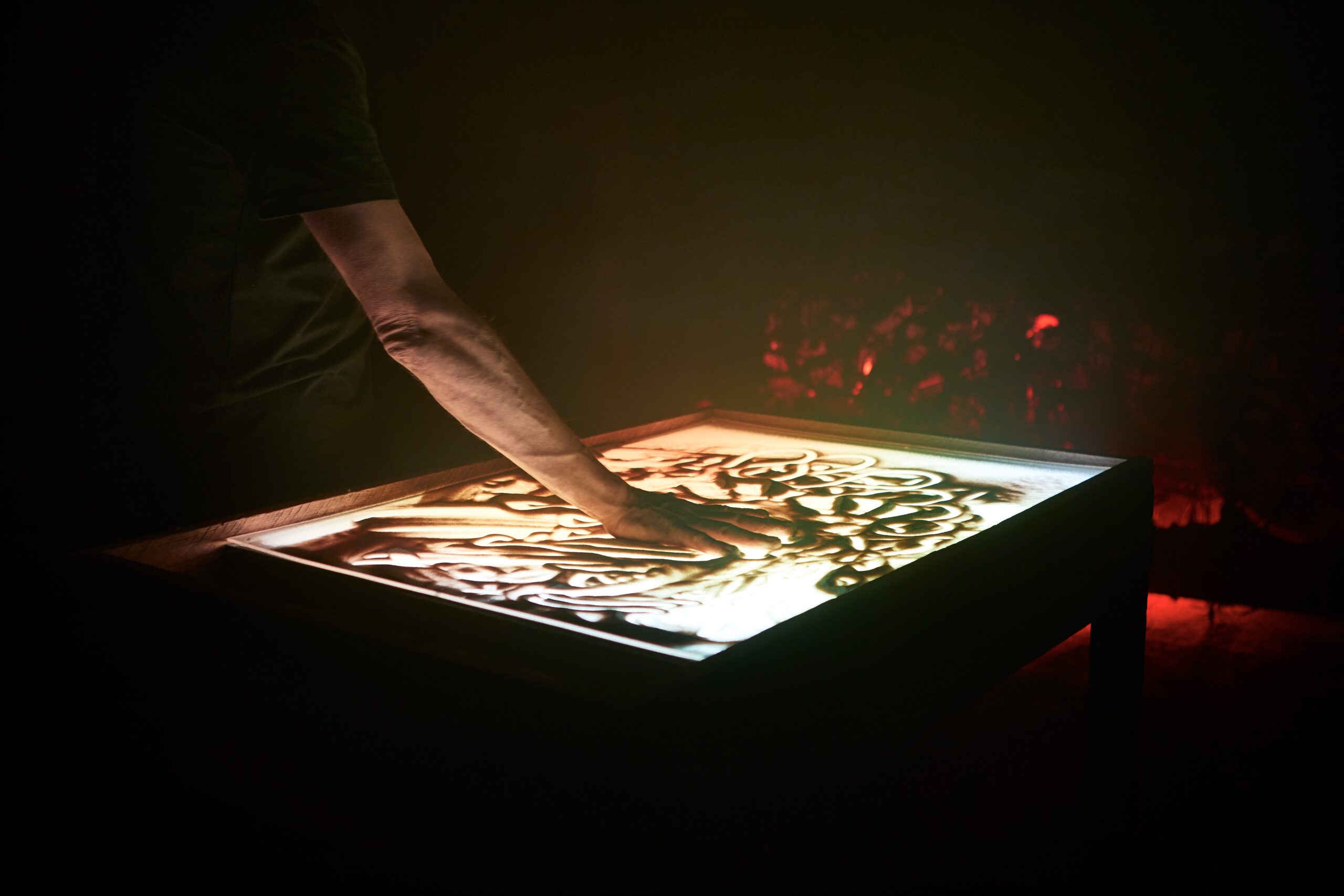 in Arm zeichnet mit dem Finger Linien in eine beleuchtete Sandfläche (Lichttisch), im dunklen Hintergrund verschwommene rote Lichtpunkte.