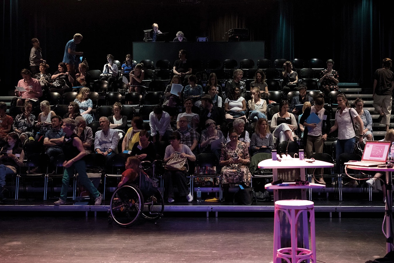 Publikum auf ansteigenden Sitzreihen in einem Theatersaal, darunter eine Person im Rollwuhl vorne am Bühnenrand. Im Vordergrund ein pink beleuchteter Hocker und ein Stehtisch mit Mikrofonen.