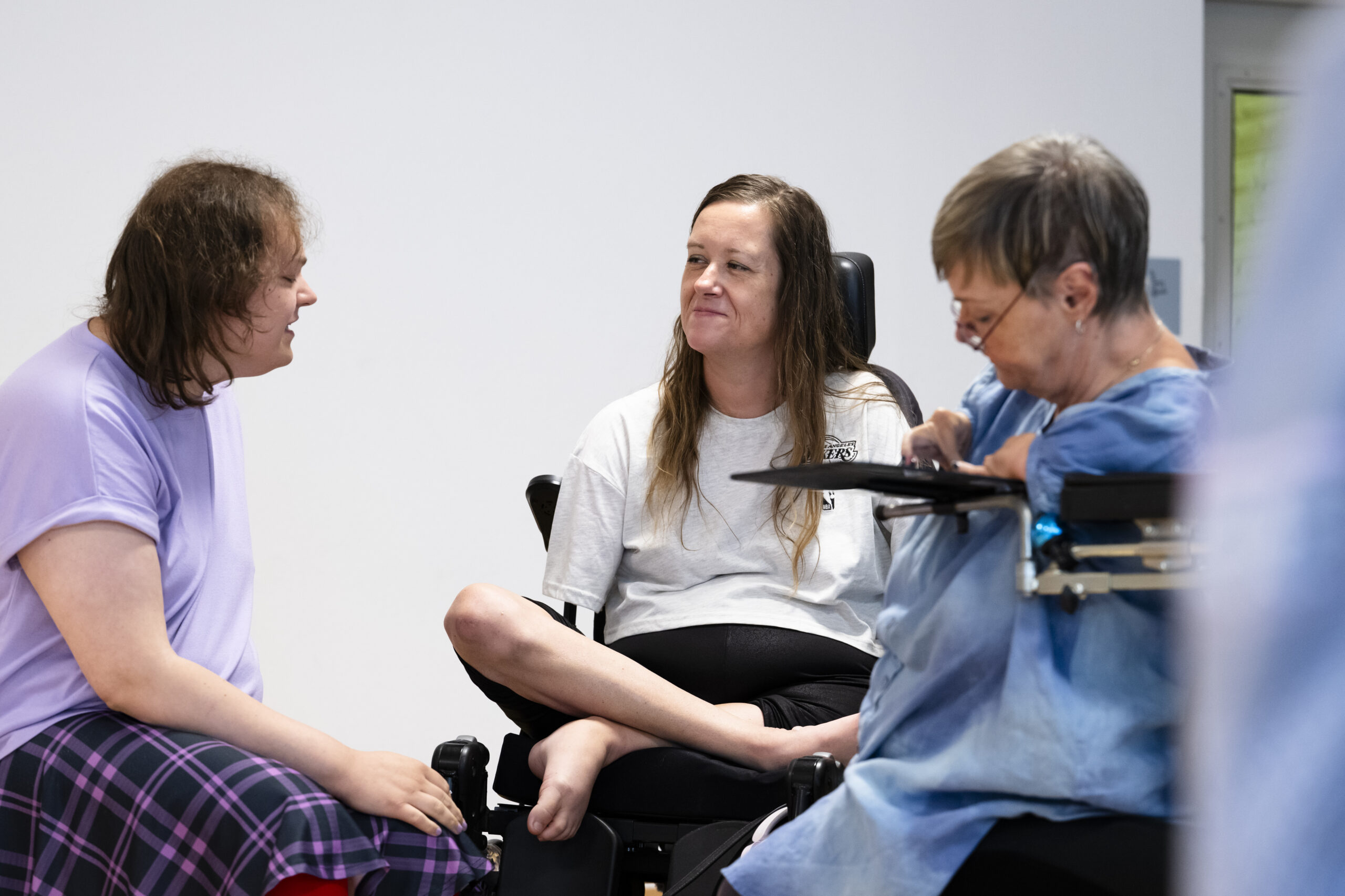 Drei Personen sitzen nah beieinander vor einer hellen Wand. Links spricht eine Person mit dunklen Haaren und lila Oberteil. In der Mitte sitzt eine Person mit langen Haaren und hellem Shirt im Elektrorollstuhl, die Beine überkreuzt, und hört lächelnd zu. Rechts im Vordergrund sitzt eine Person mit kurzen grauen Haaren und Brille in einem Rollstuhl mit angebrachtem Tablett und schaut nach unten.