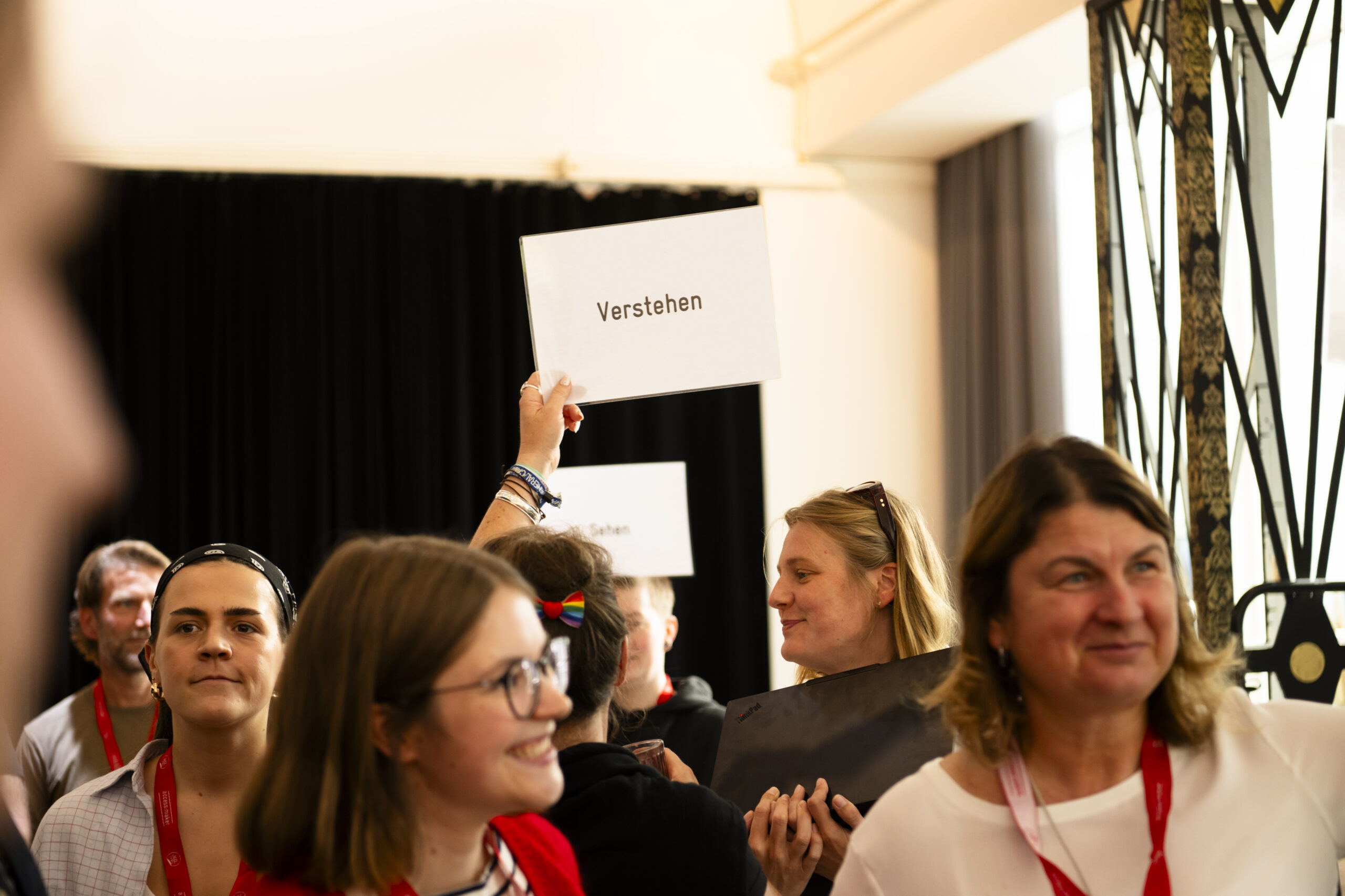 Mehrere Menschen mit roten Umhängebändern stehen dicht beieinander, einige lächeln. Im Hintergrund hält eine Person mit einem Laptop unter dem Arm ein Schild mit dem Wort „Verstehen“ hoch. Dahinter ist ein zweites Schild mit „Sehen“ zu erkennen.