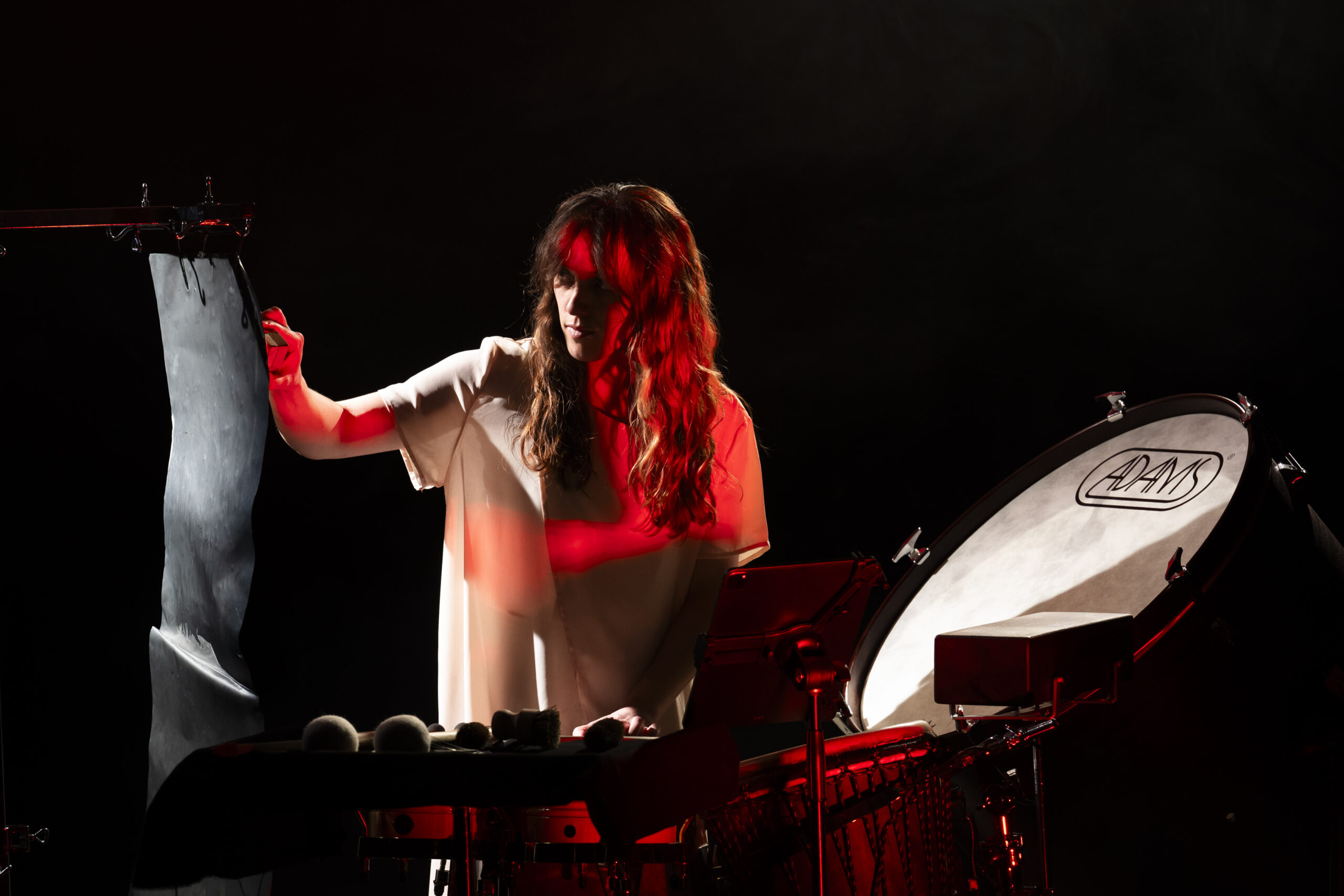 Percussionist Rita Couto Souares, with long, wavy hair and wearing a white nightdress, is playing the drums. The stage is completely dark; she is illuminated only by a red light. To the right is a large drum; to the left, a hanging sheet of metal. 