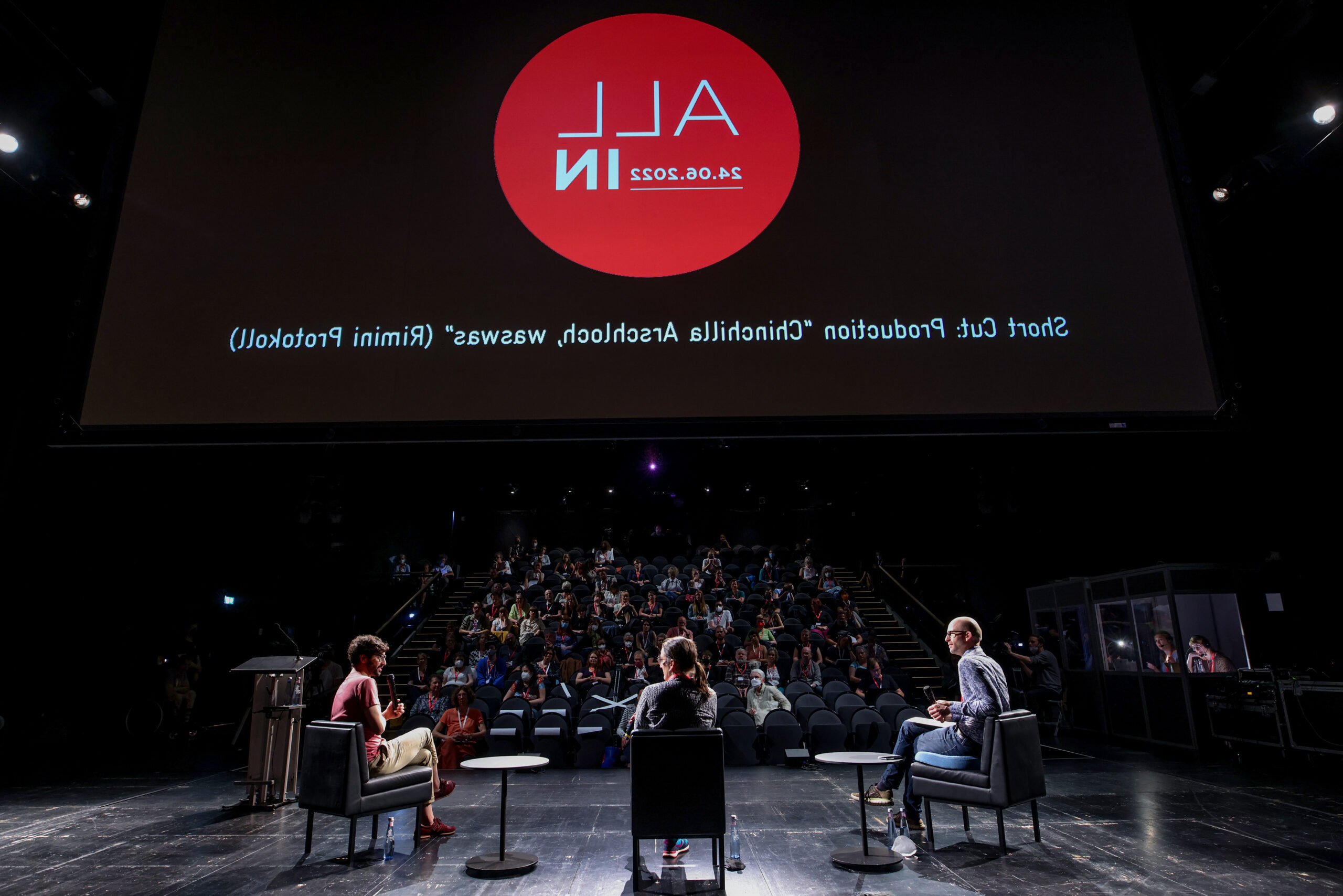 Bild vom ALL IN Symposium. Drei Personen sitzen in einer Paneldiskussion auf der Bühne, im Hintergrund sieht man das Publikum. Das Bild ist von hinter der Bühne ausgenommen.